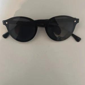Svarta runda solglasögon - Snygga svarta solglasögon med rund form och klassisk design. Bågarna är helsvarta och har små silvriga detaljer vid sidorna. Perfekta för en stilren look.