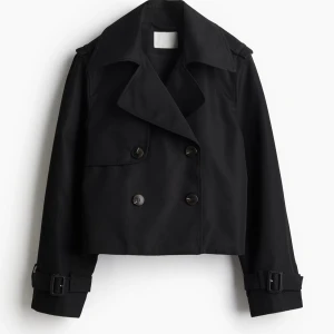 Mörkblå trenchcoat - Säljer en mörkblå kort trenchcoat med dubbelknäppning och bred krage. Jackan har bältesdetaljer vid ärmsluten och axelklaffar för en extra stilren look. Perfekt för dig som gillar trendiga och klassiska plagg. Ser nästan svart ut. Köpt för 449kr