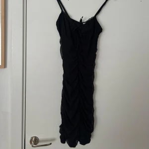 Svart tight kortklänning med smala axelband - Säljer en svart tight kortklänning med smala axelband och rynkade detaljer längs hela klänningen. Perfekt för dig som gillar en enkel men snygg look. Klänningen har en figurnära passform och är ärmlös.