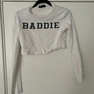 Vit croppad långärmad tröja med text - Säljer en vit croppad långärmad tröja från Bik Bok med svart text 'BADDIE' över bröstet. Perfekt till jeans eller kjol för en cool och avslappnad look.