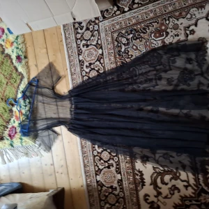 Svart långklänning i mesh med blommigt mönster - Säljer en svart långklänning i transparent mesh med blommiga broderier. Klänningen har korta ärmar och markerad midja, perfekt för dig som vill sticka ut med en unik och elegant look.