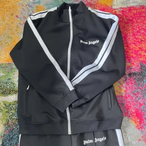 Svart träningsjacka från Palm Angels - Säljer en svart träningsjacka från Palm Angels med vita ränder längs ärmarna och dragkedja framtill. Jackan har broderad logga på bröstet och ribbade muddar. Tillverkad i polyester för en sportig känsla.