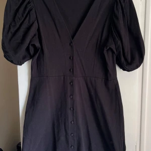 Svart klänning med puffärmar från H&M - Svart klänning från H&M med v-ringning, puffiga korta ärmar och dekorativa knappar framtill. Klänningen har en figurnära passform upptill och är sydd i ett mjukt material. Perfekt för dig som gillar stilrena och trendiga plagg. Aldrig använd