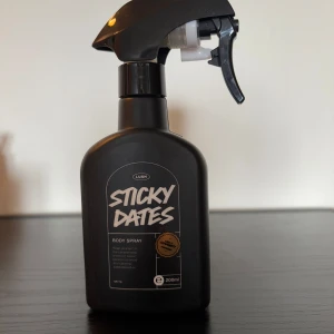 Sticky Dates Body Spray från Lush - En svart sprayflaska med doften Sticky Dates från Lush. Flaskan rymmer 200 ml och har en modern, matt finish med vit text på etiketten. Perfekt för dig som gillar söta och unika dofter. RESERVERAD