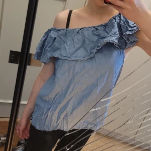 Blå jeans offshoulder volangtopp - Säljer en ljusblå jeans offshoulder topp med volangdetalj längs överdelen. Toppen är ärmlös och har en luftig passform, perfekt för varma dagar. Snygg att matcha med både jeans och kjol! Så himla mycket och trendig nu i sommar!