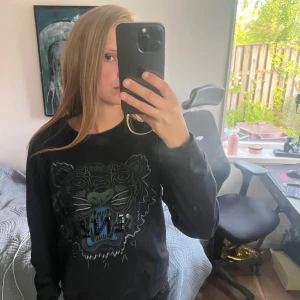 Svart sweatshirt med tiger från Kenzo - Säljer en svart sweatshirt från Kenzo med stort broderat tigerhuvud i blått, grönt och grått på bröstet. Tröjan har rund halsringning och långa ärmar. Perfekt för dig som gillar streetwear och vill sticka ut med en ikonisk design.