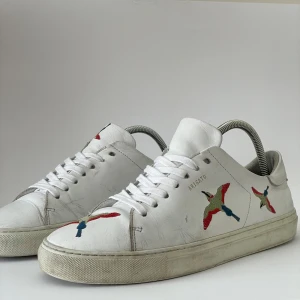 Vita sneakers från Axel Arigato🪽 - Säljer ett par vita sneakers från Axel Arigato med färgglada broderade fåglar på sidorna. Skorna har snörning, rund tå och platt sula. Perfekta för dig som gillar stilrena men unika detaljer. Original boxen kommer ej med men kommer frf med en Arigato box