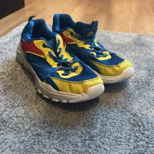 Färgglada sneakers från Lidl - Säljer ett par ikoniska Lidl-sneakers i blått, gult, rött och vitt. Skorna har en sportig design med snörning och tjock vit sula. Perfekta för dig som vill sticka ut med färg och stil. Märket syns tydligt på plösen.