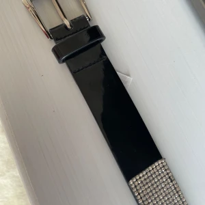 Svart glittrigt bälte från Bik Bok - Säljer ett svart bälte från Bik Bok med glittriga stenar längs hela bandet. Bältet har en klassisk silverspänne och är gjort i ett blankt material. Perfekt för att ge outfiten extra bling.