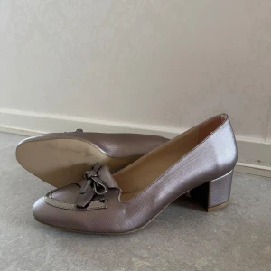 Rosa, silvriga pumps med rosett och klack - Säljer ett par eleganta silvriga pumps med låg klack och dekorativ rosett framtill. Skorna har rund tå och är tillverkade i ett glansigt syntetmaterial som ger en lyxig känsla. Perfekta för dig som vill sticka ut med stilrena detaljer. Icke använda!