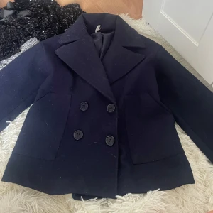 Mörkblå kappa från Design by Si - Säljer en klassisk mörkblå kappa från Design by Si. Jackan har dubbelknäppning med stora svarta knappar och bred krage. Modellen är rak och har två stora fickor framtill. Perfekt för kyligare dagar. Innan jag skickar den så tar jag bort håret ifrån jackan ❤️