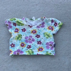 Blommig t-shirt från Name It - Söt t-shirt med färgglada blommor i lila, rosa och gult på vit botten. T-shirten har korta ärmar och rund halsringning. Perfekt för varma dagar och ger en glad sommarkänsla.