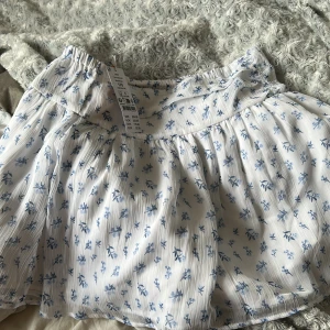 Vit blommig kortkjol från Gina  - Supersöt vit kortkjol med blått blommönster från Gina. Kjolen har låg midja och är lätt plisserad för en luftig känsla. Perfekt för varma dagar och enkel att matcha med en enkel topp. Säljer pga att den är för stor 