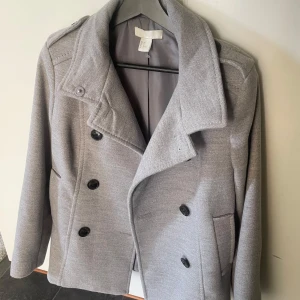 Ljusgrå kappa från H&M - Stilren ljusgrå kappa från H&M med dubbelknäppning och bred krage. Jackan har svarta knappar, axelklaffar och två fickor framtill. Perfekt för dig som vill ha en klassisk och snygg look. Använd väldigt få gånger.
