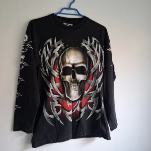 Svart långärmad tröja med dödskalletryck - Cool svart långärmad tröja med stort tryck av en dödskalle och tribal-mönster i grått och rött på både fram- och baksida. Ärmarna har även detaljerade tryck längs sidorna. Perfekt för dig som gillar en edgy stil.