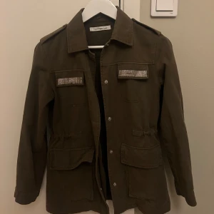 Olivgrön jacka med strassdetaljer från Copenhagen Luxe - Aldrig använd, Snygg olivgrön jacka från Copenhagen Luxe med strassdetaljer på bröstfickorna och klassisk krage. Jackan har långa ärmar, knappar framtill och stora fickor vid midjan. Vekrigen så så fin