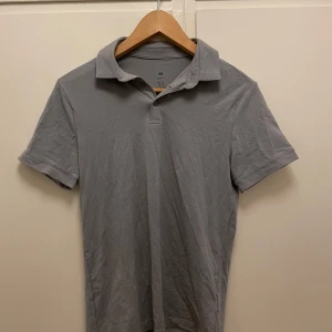 Ljusgrå pikétröja från H&M - Säljer en ljusgrå pikétröja från H&M i slim fit-modell. Tröjan har klassisk krage och korta ärmar, perfekt för en stilren och enkel look. Tillverkad i mjuk bomull för extra komfort.