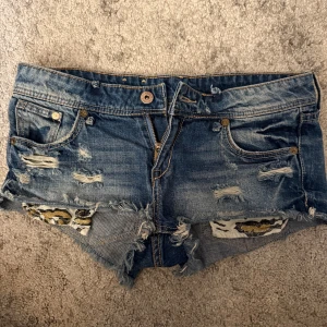 Ascoola lågmidjade shorts  - Snygga blå jeansshorts med slitna detaljer och råa kanter. Leopardmönstrad tygdetalj i guld, svart och vitt. Klassisk femficksmodell med knapp och dragkedja. Midjemått rakt över: 36,5cm🥰Priset är alltid diskuterbart