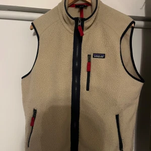 Beige fleeceväst från Patagonia - Säljer en beige fleeceväst från Patagonia med svarta detaljer och dragkedjor. Västen har en hög krage och praktiska fickor med dragkedja. Perfekt för lager-på-lager under kyligare dagar.