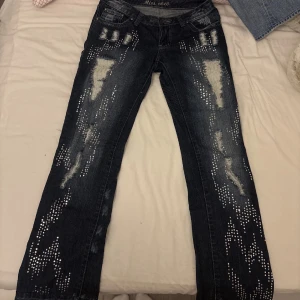 Blå bootcut jeans med slitningar och nitar - Säljer ett par blå bootcut jeans med tydliga slitningar och dekorativa nitar framtill. Jeansen har normal passform och vida ben, samt detaljerade bakfickor. De har även glitter på byxorna som är jätte fina. 💞