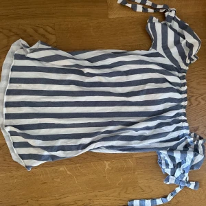 Randig offshoulder klänning från Alexander - Säljer en blåvit randig offshoulder klänning från Alexander med knytband på ena axeln. Klänningen har resår i midjan och är tillverkad i ett lätt bomullsmaterial! Använd fåtal gånger