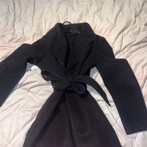 Svart kappa med knytskärp - Säljer en svart kappa med klassisk krage och knytskärp i midjan. Jackan har lång ärm och en stilren, rak passform som passar till många olika outfits. Perfekt för kyligare dagar under höst och vinter. 