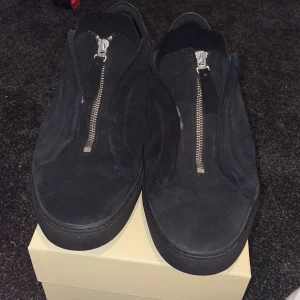 Svarta sneakers med dragkedja från Axel Arigato - Säljer ett par svarta sneakers från Axel Arigato i mocka med dragkedja framtill. Skorna har en rund tå och platt sula, samt diskret logga i guld på sidan. Snygg minimalistisk design som passar till många stilar.