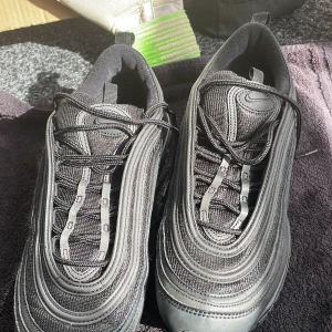 Svarta Nike Air Max 97 sneakers - Säljer ett par svarta Nike Air Max 97 sneakers med klassiska vågformade linjer och synlig luftbubbla i sulan. Skorna har snörning och är tillverkade i en mix av syntet och mesh för bra andningsförmåga. Perfekta för dig som gillar streetwear och sportig stil.