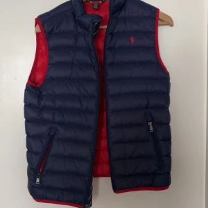 Dunväst från Polo Ralph Lauren - Säljer en mörkblå dunväst från Polo Ralph Lauren med röda detaljer och dragkedjefickor. Västen har röd insida och klassisk broderad logga på bröstet. Perfekt för lager-på-lager-stil. Pris är ej hugget i sten