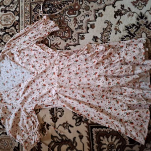 Blommig omlottklänning med volang - Söt kortklänning i ljus färg med små blommor i rosa och grönt. Klänningen har omlottmodell, volangdetaljer och korta puffärmar. Perfekt för sommarens alla tillfällen.