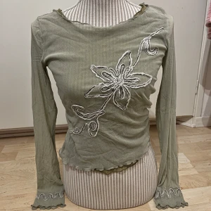Vintage topp  - Säljer en olivgrön långärmad topp med vågig kant vid hals, ärmslut och nederkant. Framsidan har en stor broderad blomma i vitt. 