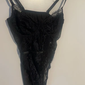 Svart spetsig bodysuit - Säljer en svart bodysuit med tunna axelband och vacker spetsdetalj. Modellen är figurnära och har transparenta partier i spetsmaterial. Perfekt för att styla under en kavaj eller till festligare tillfällen.(Aldrig använd då den är för liten) 