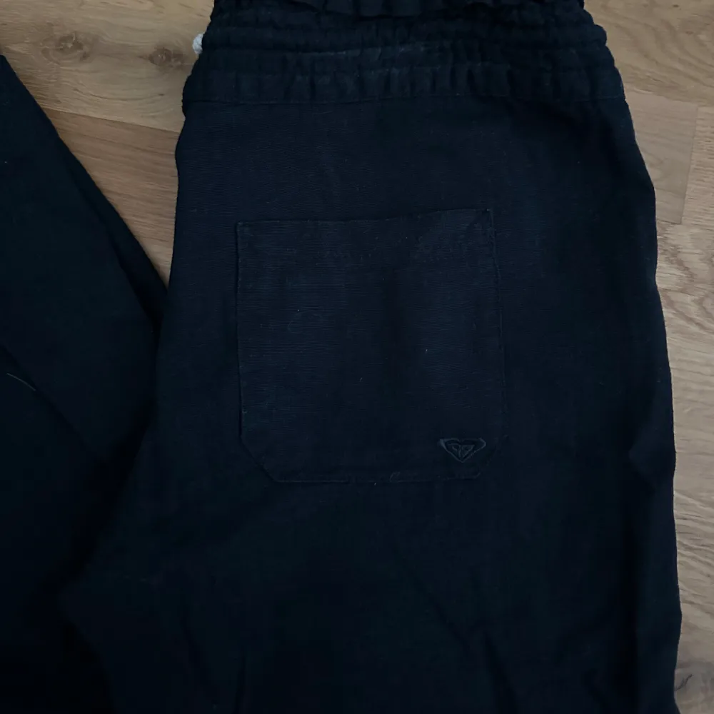 Svarta linnebyxor från Roxy med bred resår i midjan och vit snörning framtill. Byxorna har raka ben och stora fickor både fram och bak. Perfekta för en avslappnad stil.. Farkut & Housut.