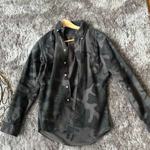 Camouflagemönstrad skjorta från Ralph Lauren - Säljer en slim fit skjorta från Ralph Lauren i grått camouflagemönster. Skjortan har långa ärmar, knappar framtill och en broderad logga på bröstet. Tillverkad i bomull. Pris går att diskutera vid snabb affär