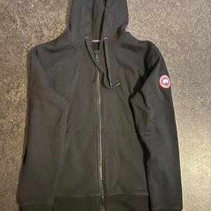  Canada Goose zip hoodie - Svart zip hoodie från Canada Goose med dragkedja framtill och huva. Klassisk logotyp-patch på vänster ärm. Jackan har en enkel och stilren design, perfekt för en avslappnad look. Tillverkad i mjukt material för extra komfort.