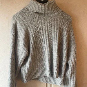 Grå croppad stickad tröja - Mysig grå stickad tröja från H&M med kabelstickat mönster framtill. Tröjan har hög polokrage och långa ärmar, perfekt för kyliga dagar. Passformen är lite oversized. Fint skick, storlek M💕