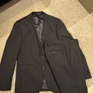 Mörkgrå kostym från Riley - Säljer en stilren mörkgrå kostym från Riley i slim fit-modell. Kavajen har två knappar, klassiska fickor och matchande byxor. Perfekt för dig som vill ha en tidlös och elegant look.