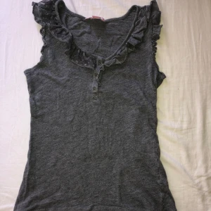 Grå ärmlös topp - Säljer en grå ärmlös topp från edc med volangdetaljer längs ringningen och knappar framtill. 