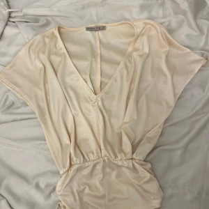 Beige v-ringad topp från Anna Field - Snygg beige topp från Anna Field med v-ringning och lös passform. Toppen har korta ärmar och resår i midjan som ger en fin siluett. Perfekt att styla till både jeans och kjol.