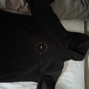 Svart hoodie från Tommy Hilfiger - Säljer en svart hoodie från Tommy Hilfiger med logga i vitt och rött framtill. Tröjan har huva och är tillverkad i bomull. Perfekt för en avslappnad stil och enkel att matcha med det mesta. I nyskick och nypris ligger runt 1.2