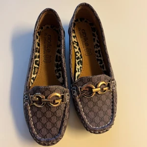 Grå loafers med gulddetaljer, ganska stora i storleken - Snygga grå loafers från Corlike med guldfärgade metallspännen framtill och diskret mönster på ovandelen. Skorna har rund tå, platt sula och ett coolt leopardmönstrat innerfoder. Perfekta för dig som vill ha en stilren men trendig look.