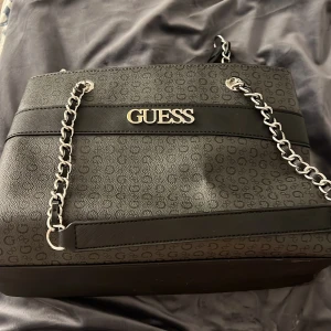 Svart axelväska från Guess med kedjerem - Säljer en svart axelväska från Guess med silverfärgad kedjerem och logotyp framtill. Väskan har präglat mönster med små G:n, flera fack inuti och stängs med magnetknapp. Perfekt för dig som vill ha en stilren och rymlig väska.