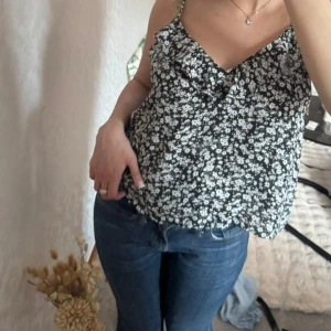 Blommigt linne med volang - Blommigt linne med vita småblommor och volangdetalj vid ringningen. Toppen har smala axelband och en luftig passform. Storlek L men är liten i storlek!🌸