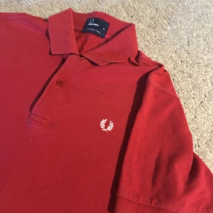 Fred Perry pike - Säljer en klassisk röd piké t-shirt från Fred Perry med vit broderad logga på bröstet. T-shirten har krage, knappar vid halsen och är tillverkad i 100% bomull. Perfekt för en stilren och sportig look.