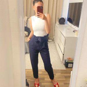 Mörkblå randiga kostymbyxor med knytband - Säljer ett par mörkblå kostymbyxor med tunna vita ränder och knytband i midjan. Byxorna är högmidjade och har en rak passform som ger en stilren look. Perfekta att styla med en enkel topp eller skjorta.