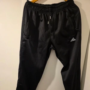 Svarta träningsbyxor från Adidas - Svarta träningsbyxor från Adidas med resår i midjan och dragkedjor vid fickorna. Byxorna har en sportig look med Adidas-logga på ena benet och är perfekta för träning eller chill.  Storlek L **oanvänd köpt av en loppis