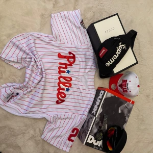 Vit Phillies baseballtröja med röda ränder - Säljer en vit baseballtröja från Majestic med röda ränder och Phillies-logga framtill. Nummer 28 och namnet Werth på ryggen. Knappar framtill och korta ärmar. Perfekt för dig som gillar sportig stil.