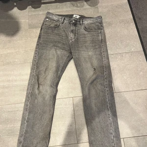 Grå raka jeans - Säljer ett par grå jeans med rak passform och klassisk femficksdesign. Jeansen har en diskret sliten detalj på ena benet och stängs med dragkedja och knapp. Perfekta för en avslappnad stil.