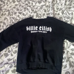 Svart sweatshirt Billie Eilish 'Happier Than Ever' - Svart sweatshirt med vit text 'billie eilish happier than ever' och ett tryck av Billie Eilish på framsidan. Tröjan har rund hals och långa ärmar. Perfekt för fans av Billie Eilish.