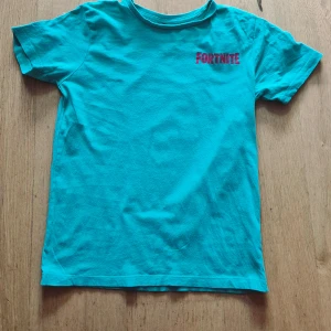 Turkos Fortnite t-shirt med taco-tryck - Säljer en grön t-shirt med Fortnite-logga på bröstet och ett stort, färgglatt taco-tryck på ryggen. Perfekt för dig som gillar gaming och coola motiv! T-shirten har rund hals och är kortärmad.